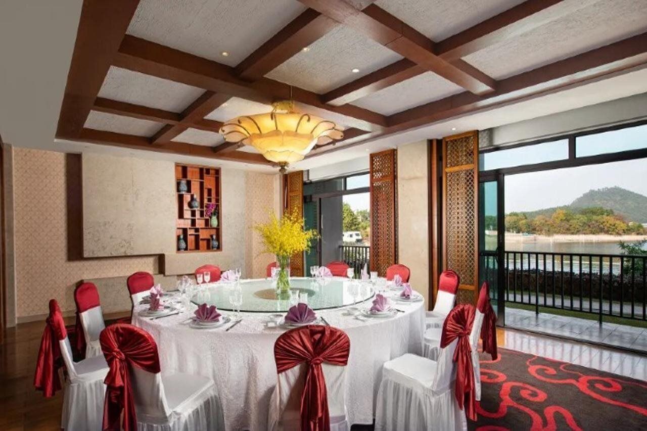 A round dining table with white tablecloth and red sashes, surrounded by chairs with red sashes. A glass-top table is set with plates, silverware, and a vase with yellow flowers. The room has a high ceiling with wooden beams, a large chandelier, and a view of a mountain through a balcony railing.