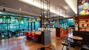 Un café amplio con suelo de madera, sillas rojas y negras, una zona de bar central y grandes ventanales que ofrecen vistas a zonas verdes.