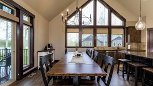 Un comedor amplio con una larga mesa de madera, múltiples sillas y una lámpara de araña. Amplias ventanas ofrecen luz natural y vistas al exterior.