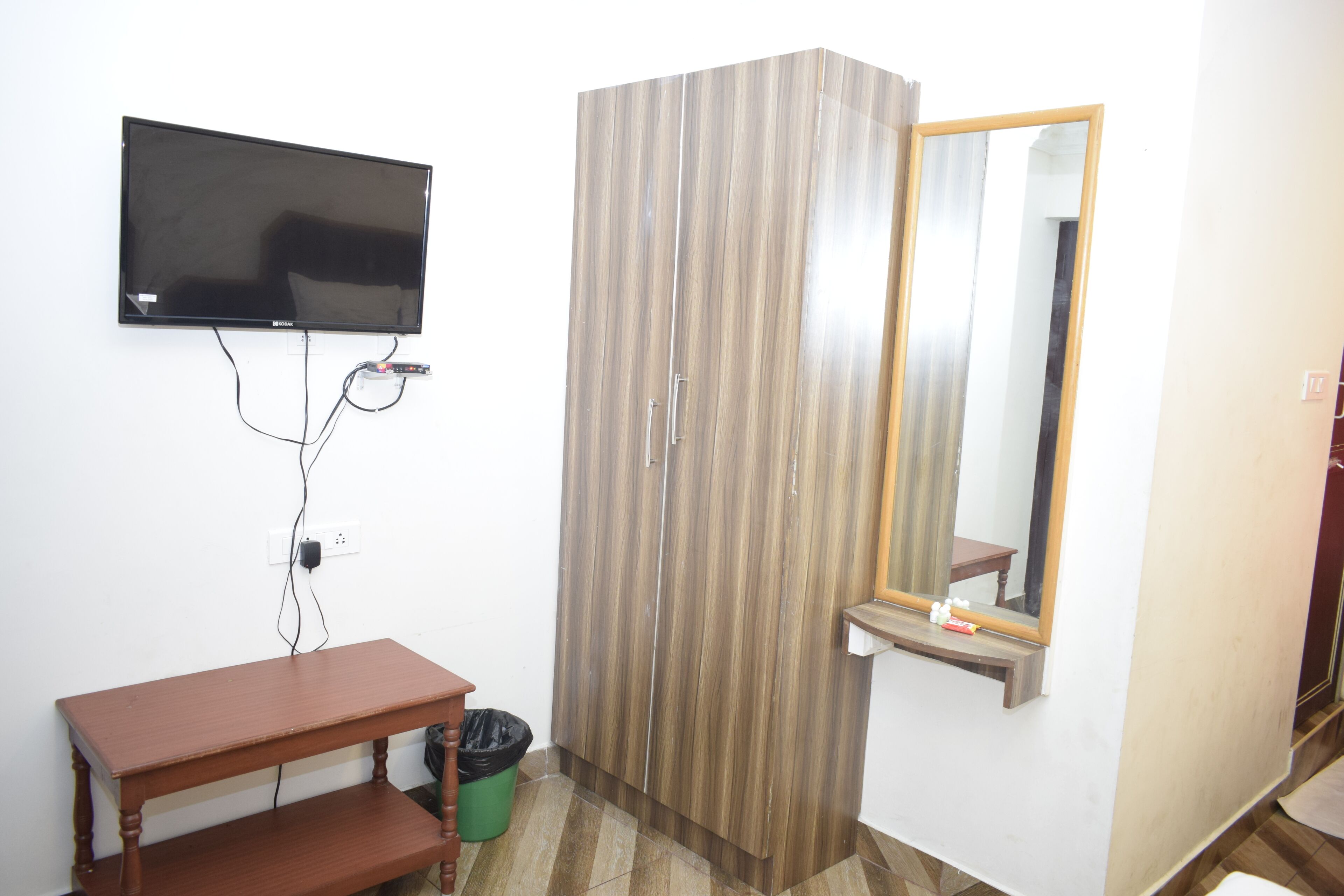 A room with a flat-screen TV mounted on the wall, a wooden wardrobe, a small wooden table, and a mirror above a sink.