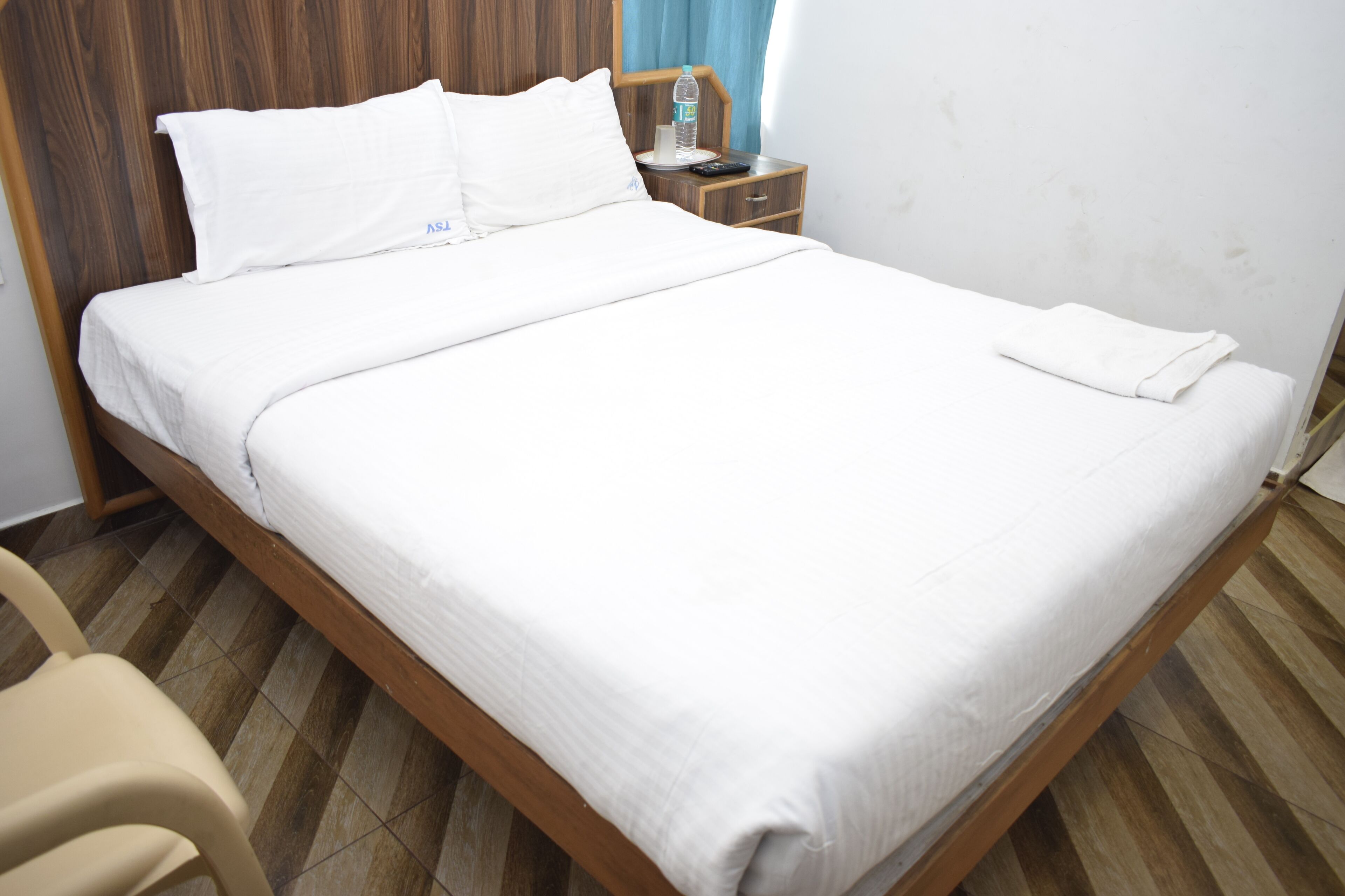 Una cama bien hecha con sábanas blancas, cabecera de madera, mesita de noche con una botella de agua y una silla.