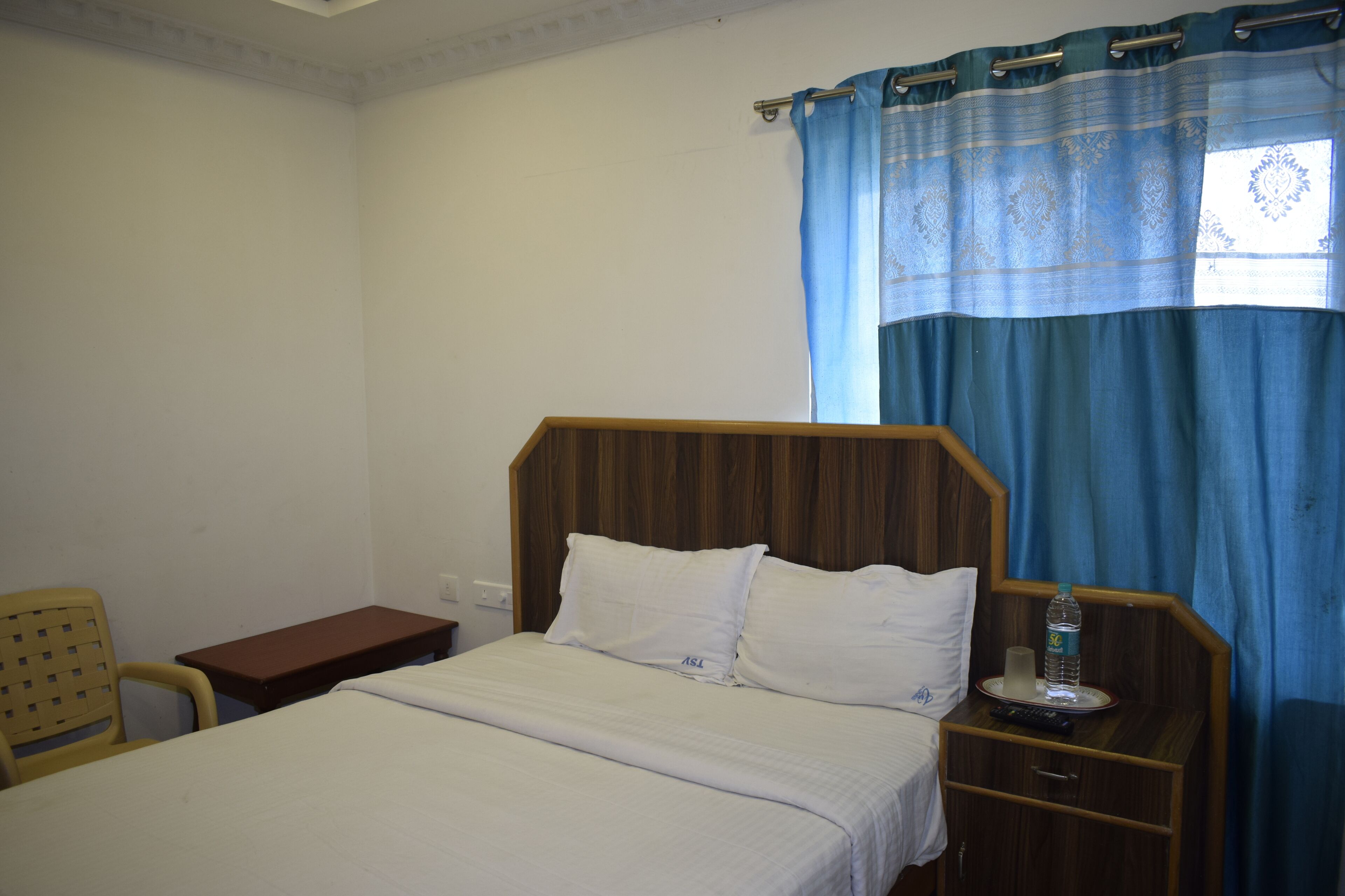 Una cama individual con ropa blanca, cabecera de madera, mesita de noche con un control remoto, una botella de agua y una pequeña mesa.