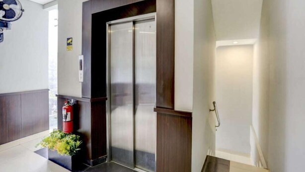 Een hotellobby met een receptiebalie, een brandblusser, een ventilator aan de muur en een lift.