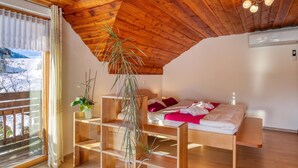 A room with a wooden ceiling, a bed with white bedding and red pillows, a wooden shelf with plants, and a window with curtains.