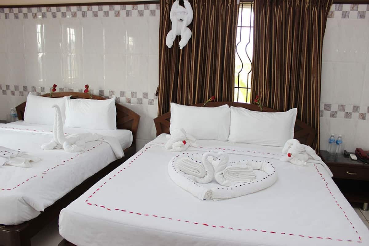 A hotel room with two beds, each with white linens and decorative swan-shaped towels. The room has a checkered tile border, a window with curtains, and a wooden nightstand with a phone and water bottles.