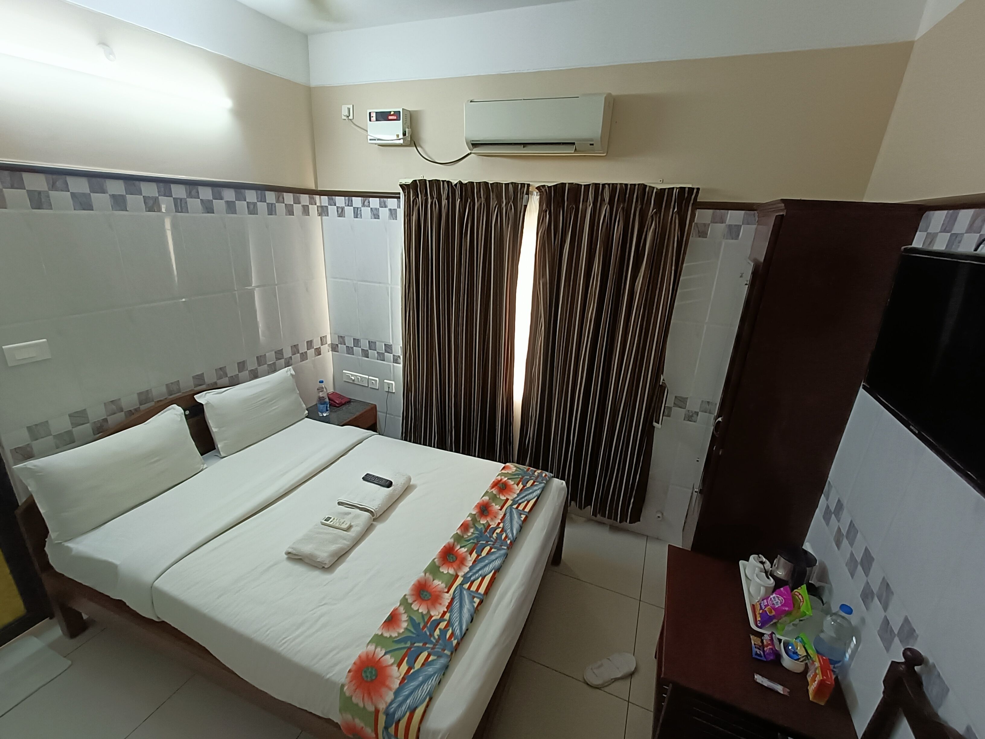A hotel room with a bed, a bedside table, a TV, an air conditioner, and a window with curtains.