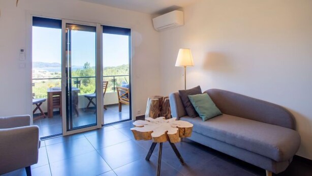 Ein modernes Wohnzimmer mit einer Couch, einem Holztisch und einer Lampe. Ein Schiebeglasfenster führt zu einem Außenbereich mit Tisch und Stühlen.