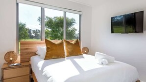 Un dormitorio moderno con un ventanal grande, una cama con ropa de cama blanca, un televisor de pantalla plana y una mesita de noche de madera con una lámpara.