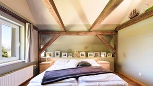 Une chambre avec un plafond en pente, des poutres en bois, un lit recouvert de draps blancs, une fenêtre avec des rideaux et des tableaux encadrés accrochés au mur.