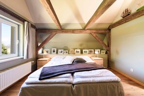 A bedroom with a sloped ceiling, wooden beams, a bed with white linens, a window with curtains, and framed pictures on the wall.