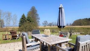 Une terrasse extérieure avec une table et des chaises en bois, un fauteuil en osier, une lampe en osier et un grand parasol.