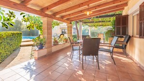 Een overdekt terras met betegelde vloer, houten balken, tuinmeubilair, potplanten en uitzicht op een zwembad.