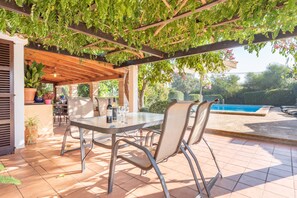 Une terrasse extérieure avec une table et des chaises, une pergola couverte de verdure, une piscine et un jardin en arrière-plan.