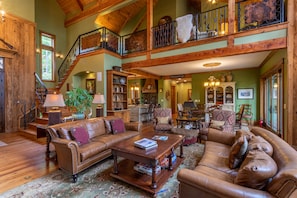 A spacious living area with a high ceiling, wooden beams, a staircase, and a large window.