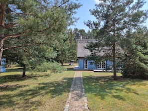 Une maison rustique avec un toit de chaume, entourée d’une dense forêt de pins et un chemin pavé menant à l’entrée.