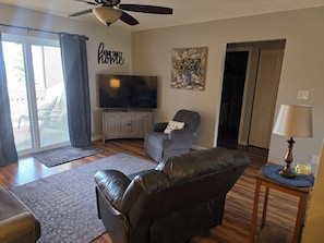 A living room with a flat-screen TV, a ceiling fan, a gray armchair, a leather sofa, a wooden coffee table, and a painting on the wall.