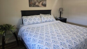 A neatly made bed with a floral-patterned bedspread, a small bedside table with a lamp, a wall-mounted painting, and a potted plant. - Lee's Helena Hideout (Helena)