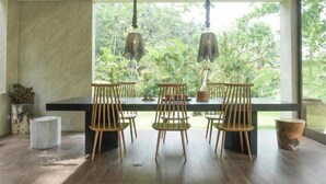 A dining area with a long table, six wooden chairs, hanging light fixtures, and a view of lush greenery through large windows. - Sandhya Villa Ubud (Payangan)