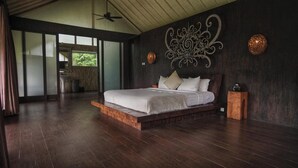 A spacious bedroom with a large bed, dark wooden flooring, and a view of a bathroom and greenery outside. - Sandhya Villa Ubud (Payangan)