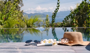 A glass of water, sunglasses, a hat, and flowers on a table by a pool with a mountain view.