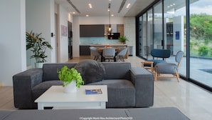 A modern living room with a grey sofa, a white coffee table, and a potted plant. There is a dining area with a table and chairs in the background. - The Bridge Luxury Villa (Nha Trang)