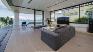 A modern living room with a large flat-screen TV, a grey sofa, and a coffee table. There is a large window offering a view of the ocean and mountains. - The Bridge Luxury Villa (Nha Trang)