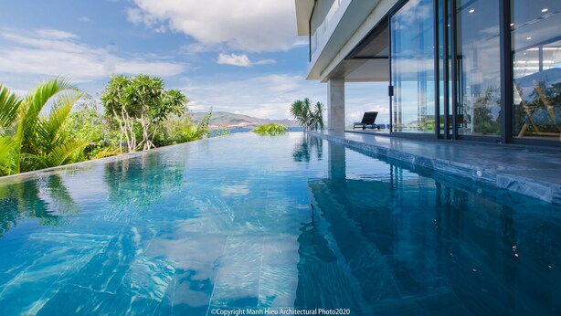 A modern infinity pool with clear blue water, surrounded by lush greenery and a view of distant hills. - The Bridge Luxury Villa (Nha Trang)