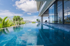 A modern infinity pool with clear blue water, surrounded by lush greenery and a view of distant hills.