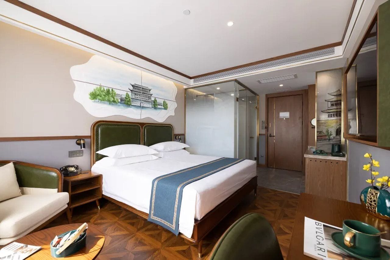 Habitación de hotel con cama, escritorio, sillón, espejo y un cuadro en la pared.