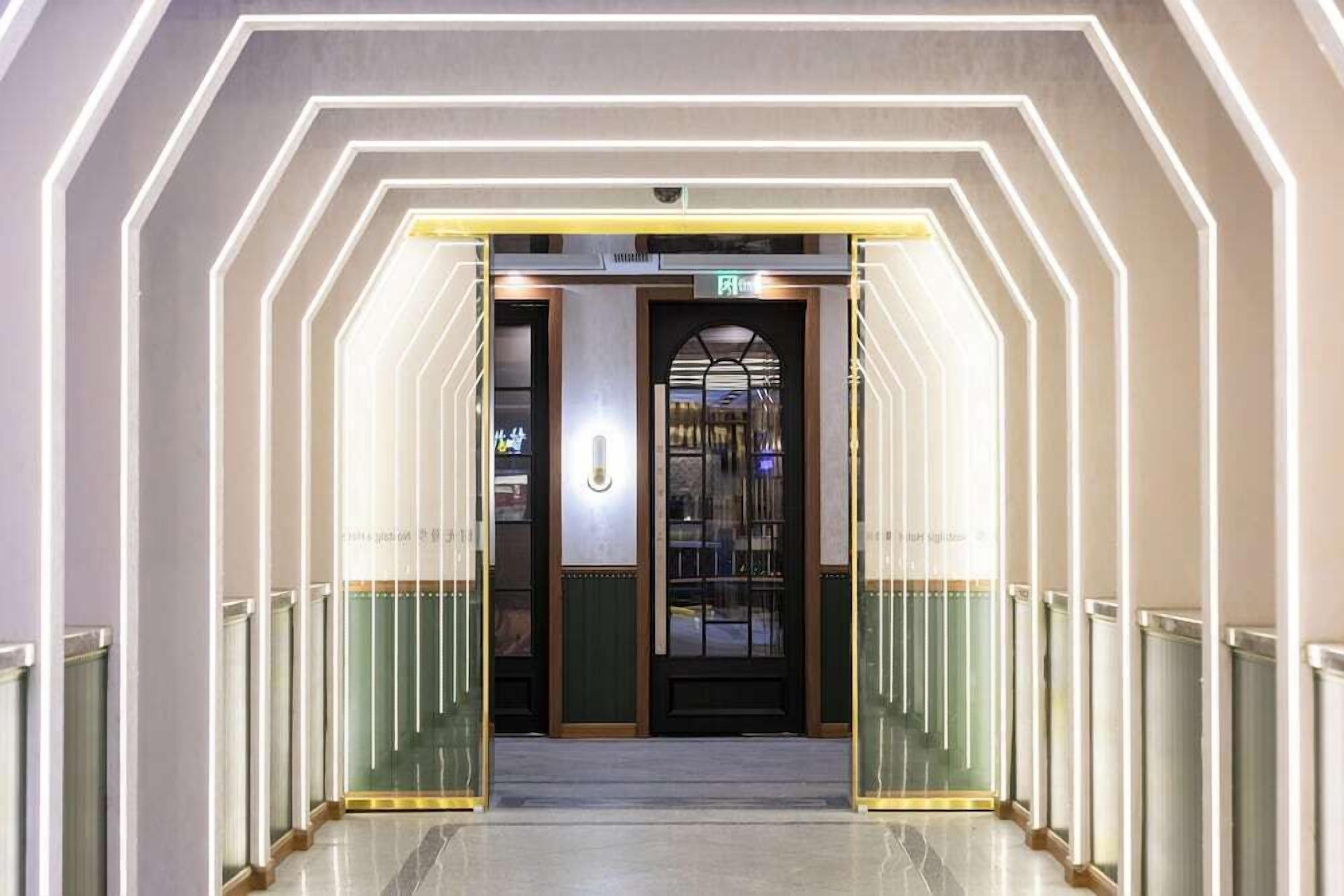 Un pasillo de hotel con un diseño de iluminación geométrico, una entrada con puerta de vidrio y una señal de salida visible.