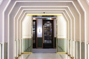 Un couloir d’hôtel avec un éclairage aux motifs géométriques, une entrée avec une porte en verre et un panneau de signalisation indiquant la sortie, bien visible.