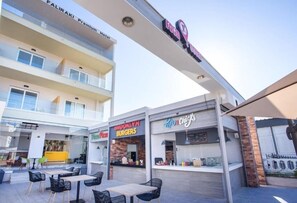 An outdoor dining area with tables and chairs, a food court with various eateries, and a building with a sign for 'FALIRAKI Premium Hotel' in the background. - Faliraki Premium Hotel (Rhodes)