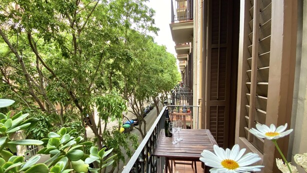 Un balcone con tavolo e sedie in legno, immerso nel verde di alberi rigogliosi e con una fila di auto parcheggiate.
