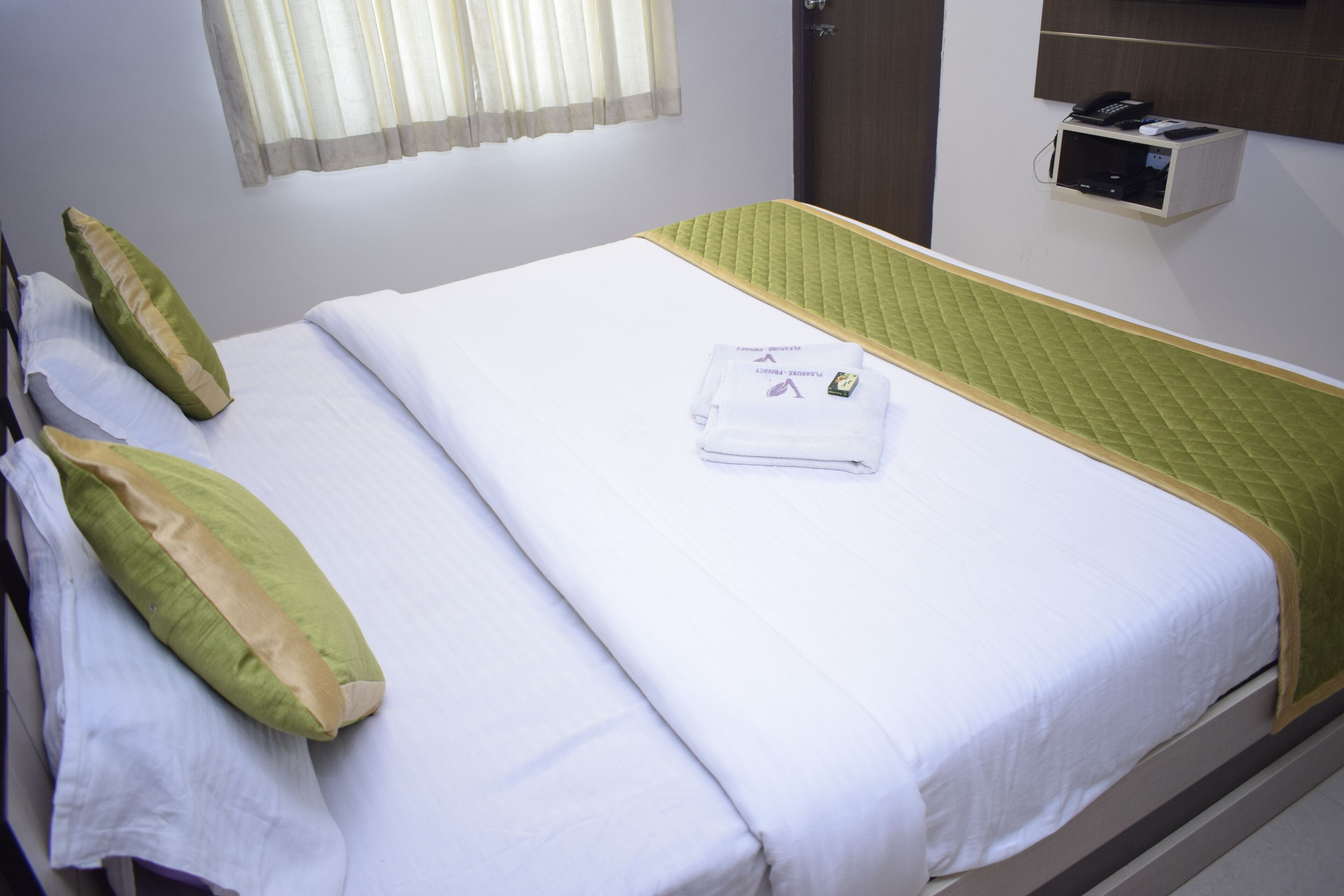 Una cama bien hecha con ropa de cama verde y blanca, una toalla doblada y un teléfono en la mesita de noche.