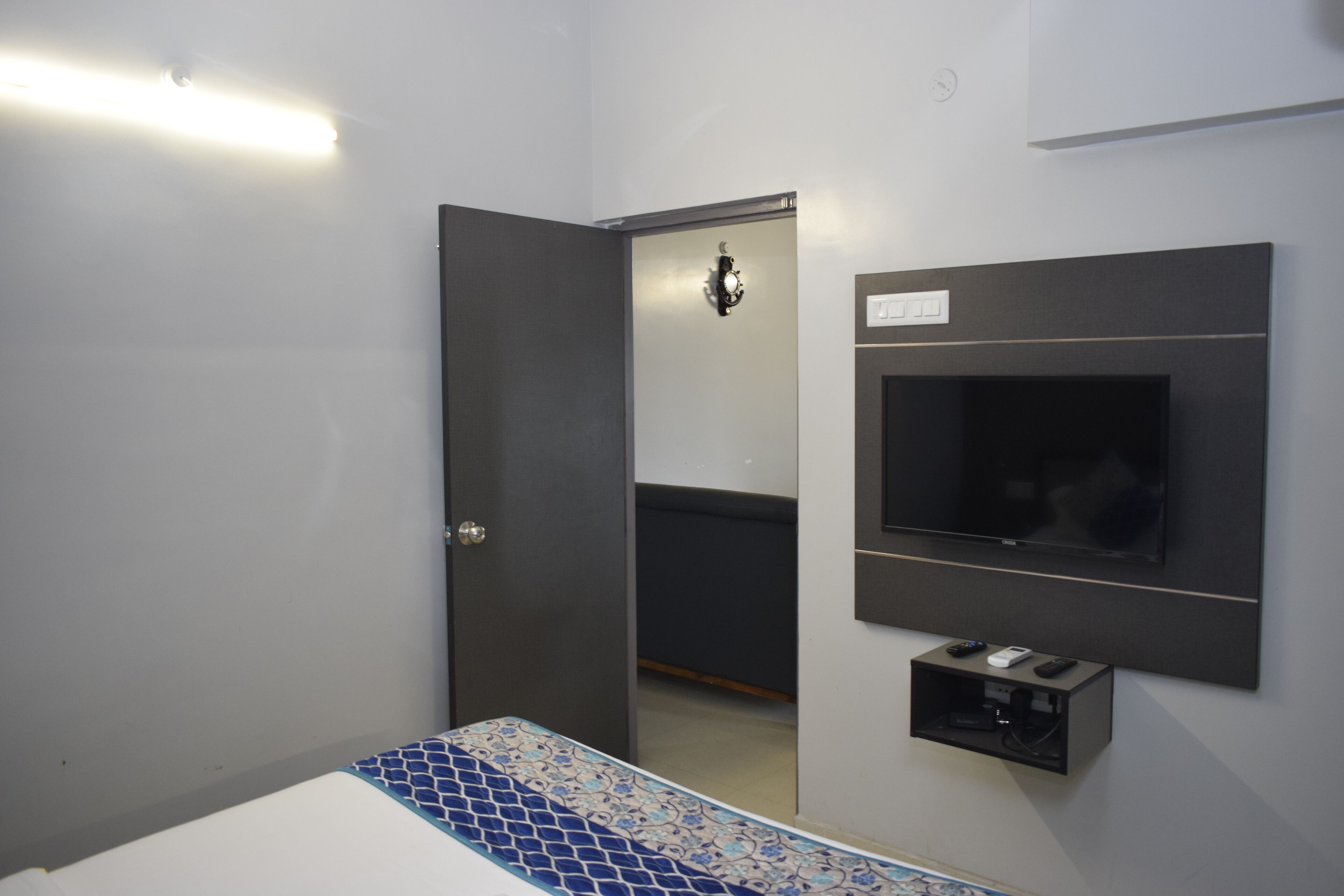 Ein Hotelzimmer mit Bett, Wandfernseher, einer kleinen Ablage für elektronische Geräte und einer Tür zu einem weiteren Raum.