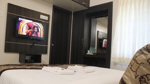 Ett hotellrum med en säng, en tv som är monterad på väggen, en spegel och ett litet bord med en flaska och ett glas.