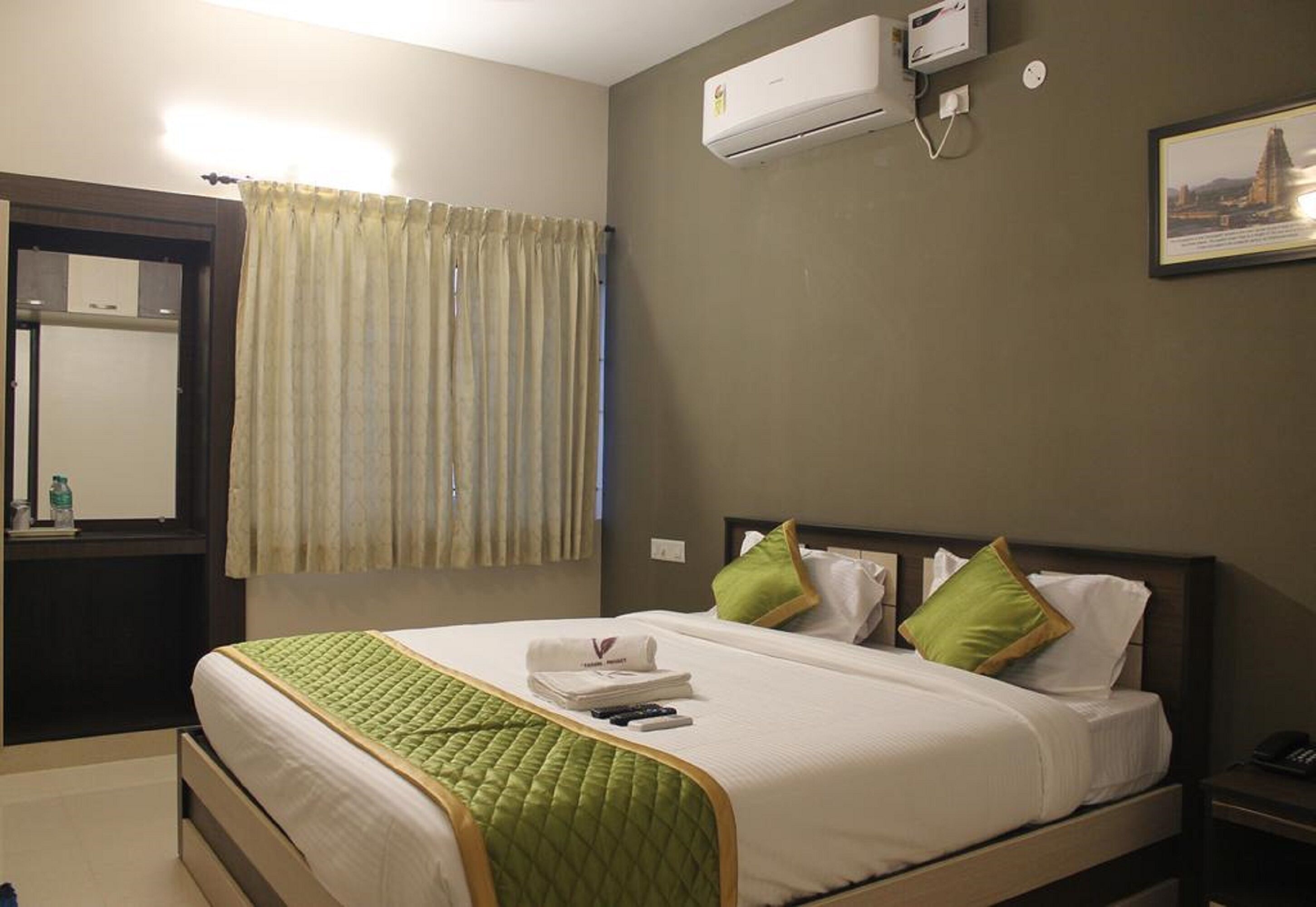A hotel room with a bed, a nightstand, an air conditioning unit, a window with curtains, and a framed picture on the wall.
