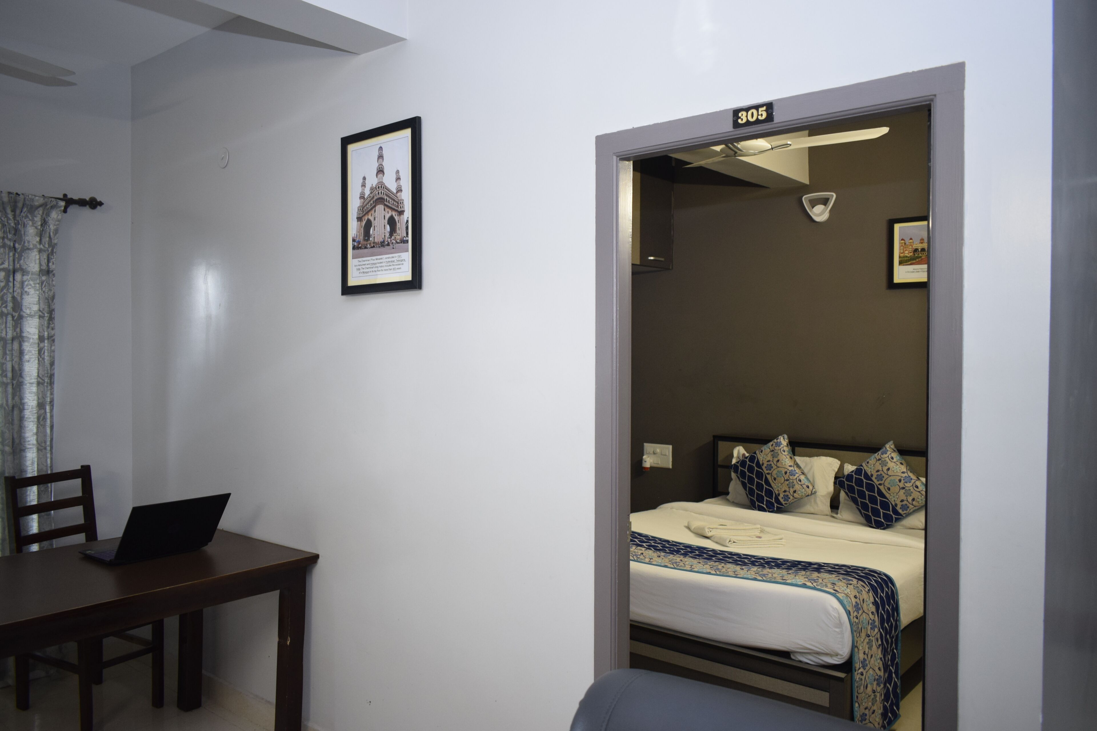 A room with a bed, a desk with a laptop, a framed picture on the wall, and a door with a number.