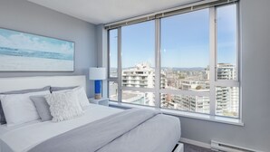 Moderni hotellihuone, jossa on suuri sänky, yöpöytä, lamppu ja ikkuna, josta avautuu näkymä kaupunkiin.