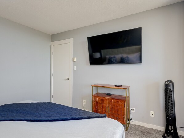 A bedroom with a bed, a TV mounted on the wall, a wooden cabinet, and a standing fan.