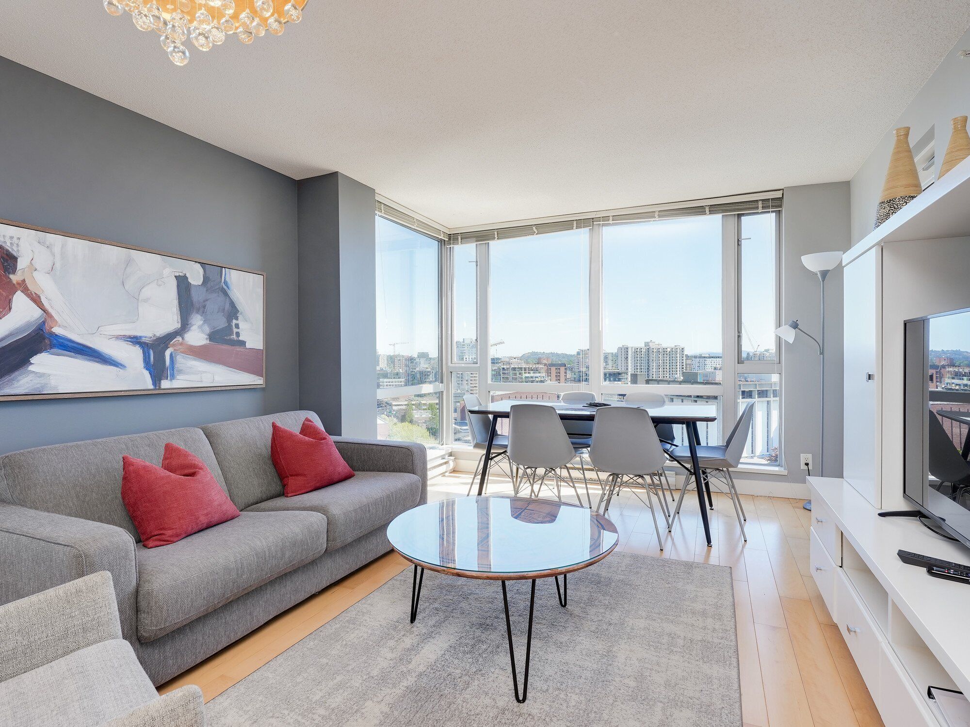 Un salon moderne avec un canapé gris, des coussins rouges, une table basse en verre et une grande fenêtre donnant sur la ville.