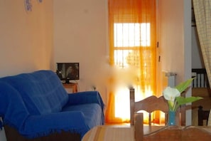Un salon avec un canapé bleu, un téléviseur posé sur un meuble et une fenêtre avec des rideaux.