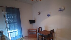 Una sala de estar con un sofá, una mesa de comedor con sillas, un televisor montado en la pared y una lámpara en el techo.