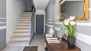 Una escalera que lleva a una puerta, una mesa de madera con libros y una planta en maceta, y un espejo en la pared.