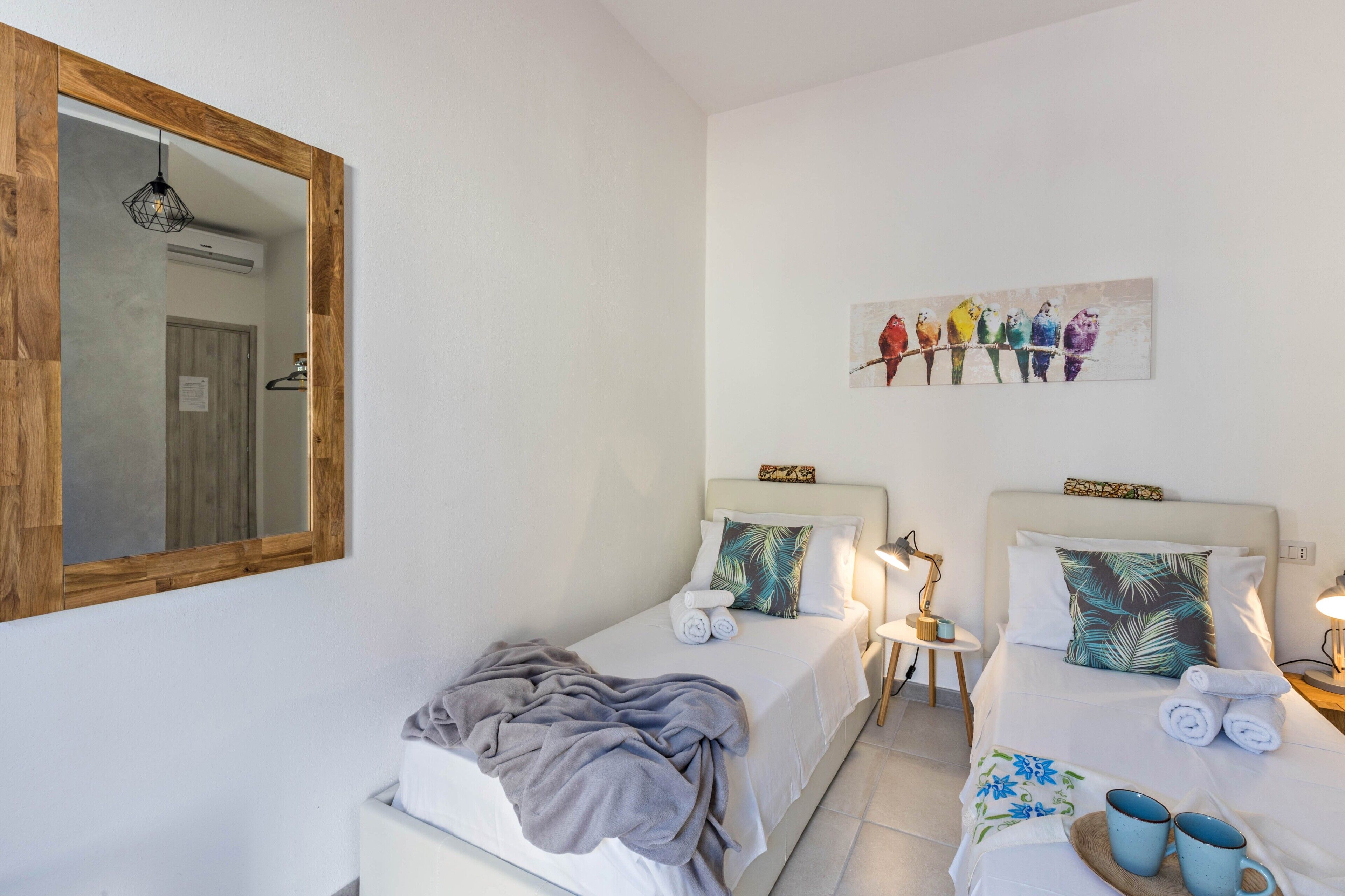 Habitación de hotel con dos camas, un espejo grande, un cuadro con aves de colores y una mesita con lámpara.