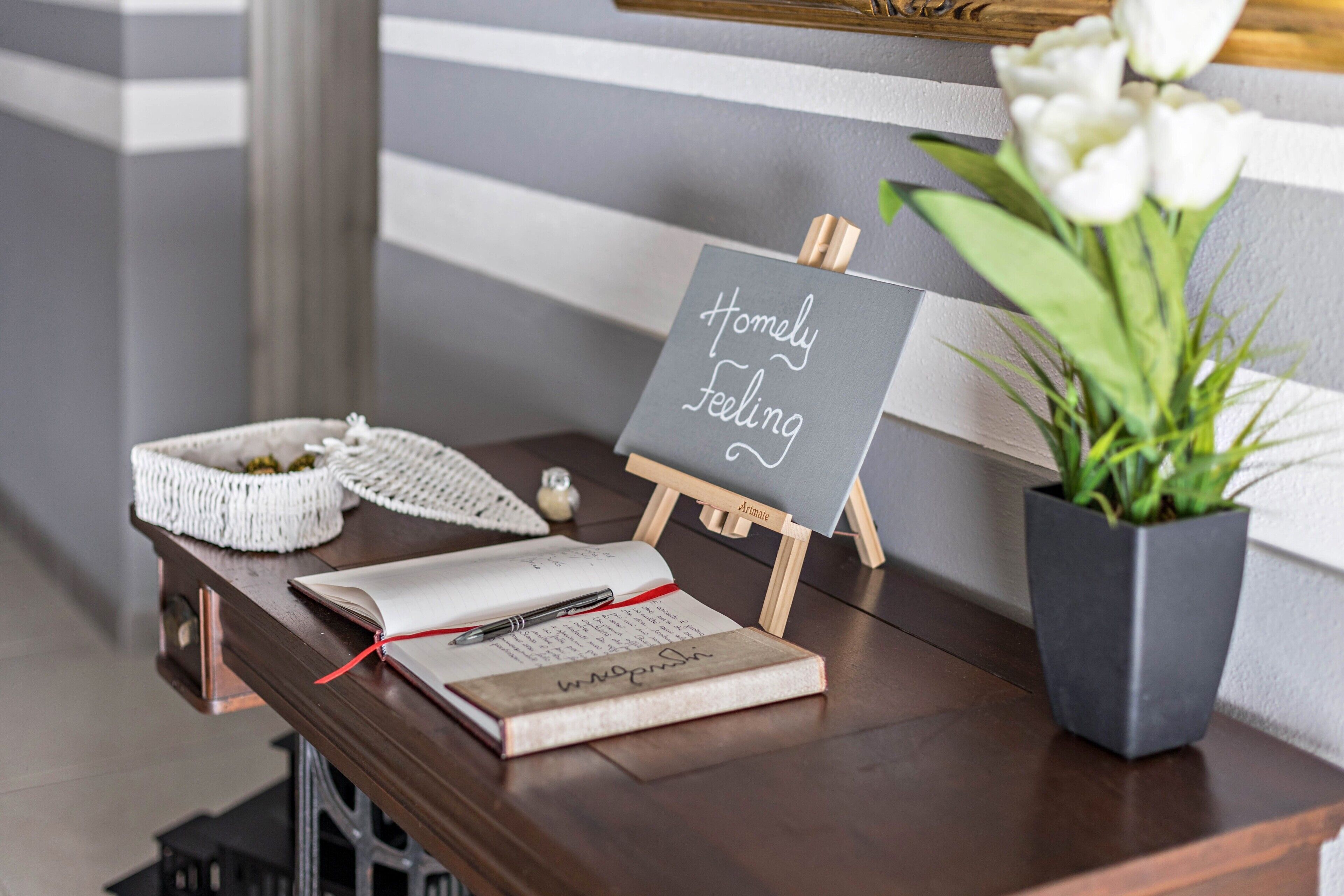 A wooden table with a notebook, a pen, a small chalkboard with 'Homely Feeling' written on it, a white crocheted basket, and a potted plant.