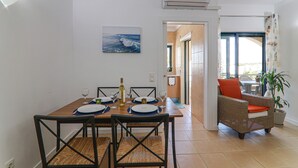 Un área de comedor con una mesa dispuesta para cuatro personas, una silla de mimbre con cojín naranja y una planta en maceta.