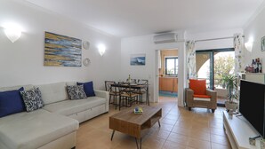 Una sala de estar con sofá, mesa de centro, mesa de comedor y sillas. Hay un televisor y una planta.
