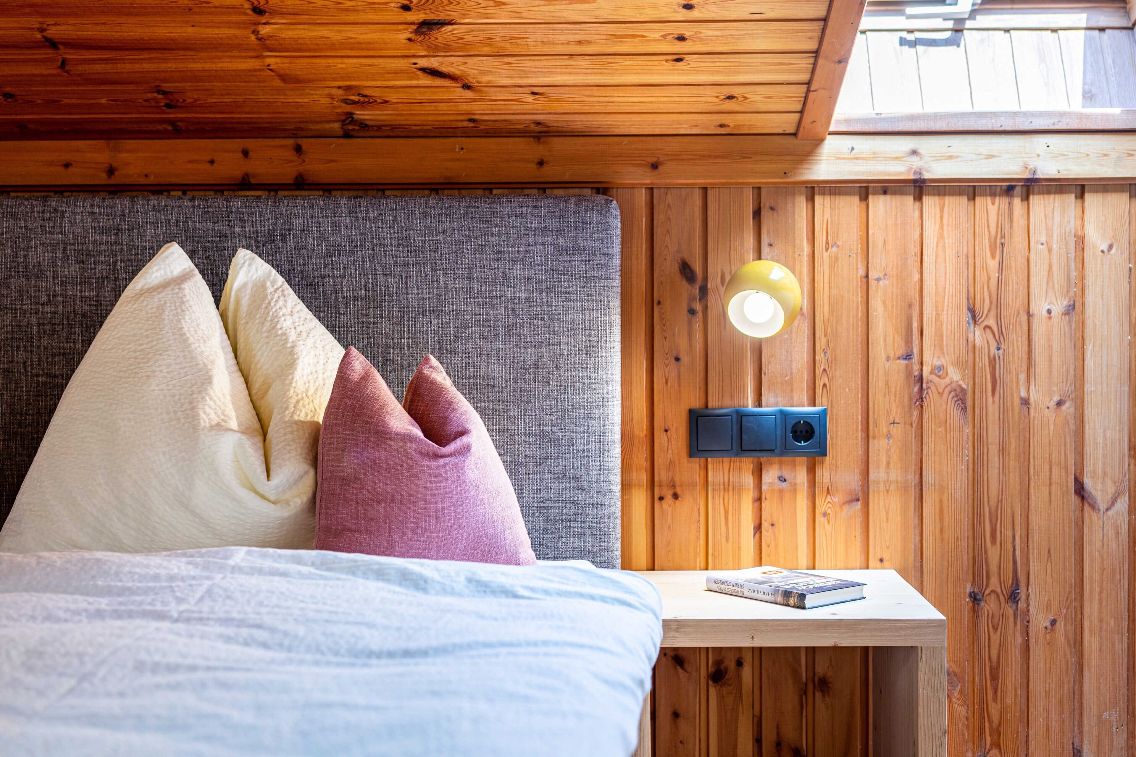A wooden-paneled room with a bed, pillows, a bedside table with a book, and a wall-mounted light fixture.