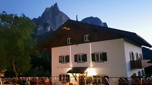 A building with a white facade, green shutters, and a balcony. It is situated in a mountainous area with a prominent peak in the background.